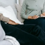 Unrecognizable professional female psychologist writing on clipboard while sitting against client on blurred background during psychotherapy session in light office