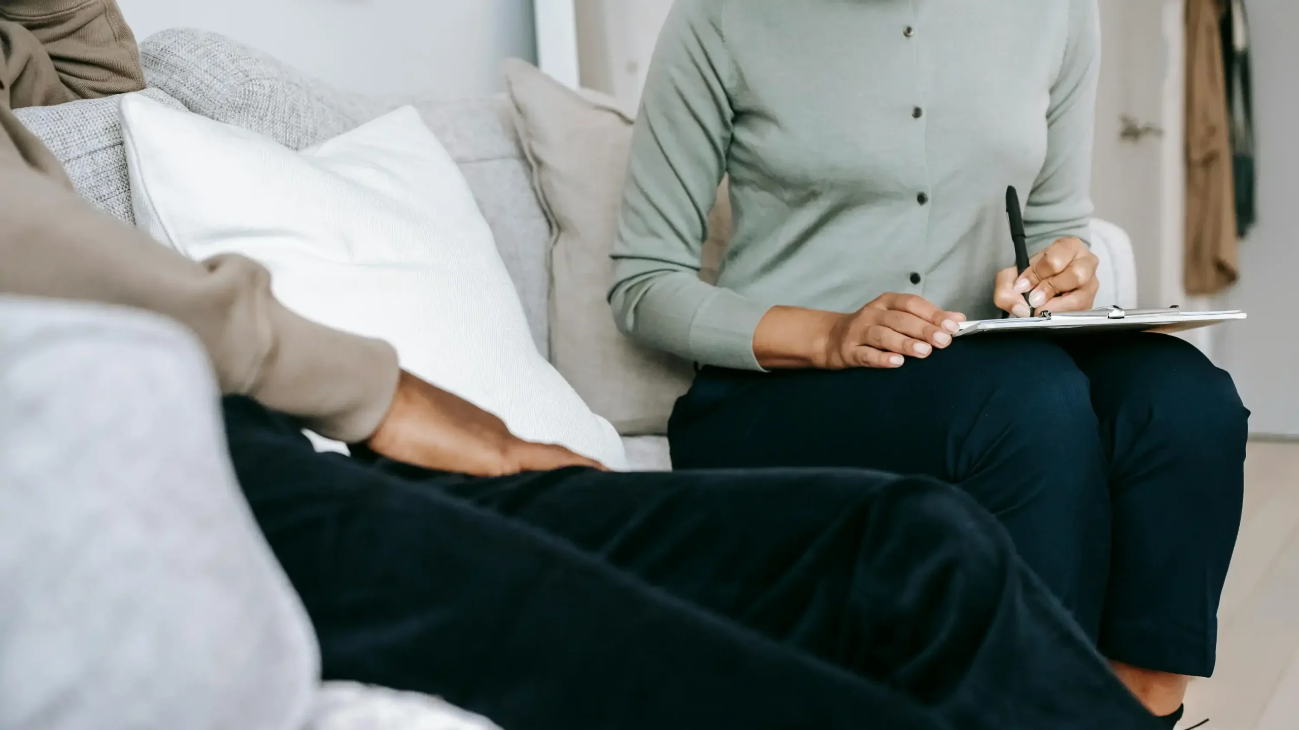 Unrecognizable professional female psychologist writing on clipboard while sitting against client on blurred background during psychotherapy session in light office